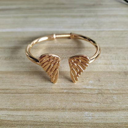 A rose gold cuff bracelet featuring two textured angel wing charms, displayed on a light wood grain surface.