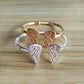 A pair of open cuff rings with textured angel wings, one in polished rose gold and the other in polished silver, displayed on a light wood background.