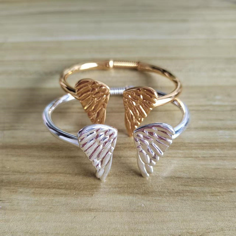 A pair of open cuff rings with textured angel wings, one in polished rose gold and the other in polished silver, displayed on a light wood background.