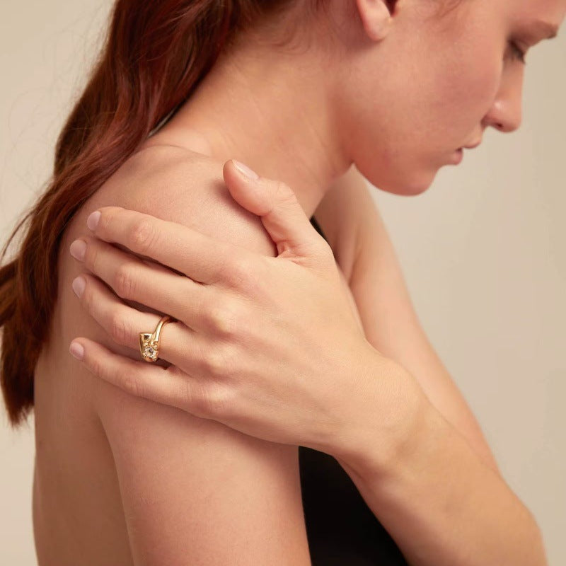 A close-up lifestyle shot of a woman wearing a gold ring with a small clear gemstone on her index finger, holding her shoulder. The background is a solid, warm beige tone.