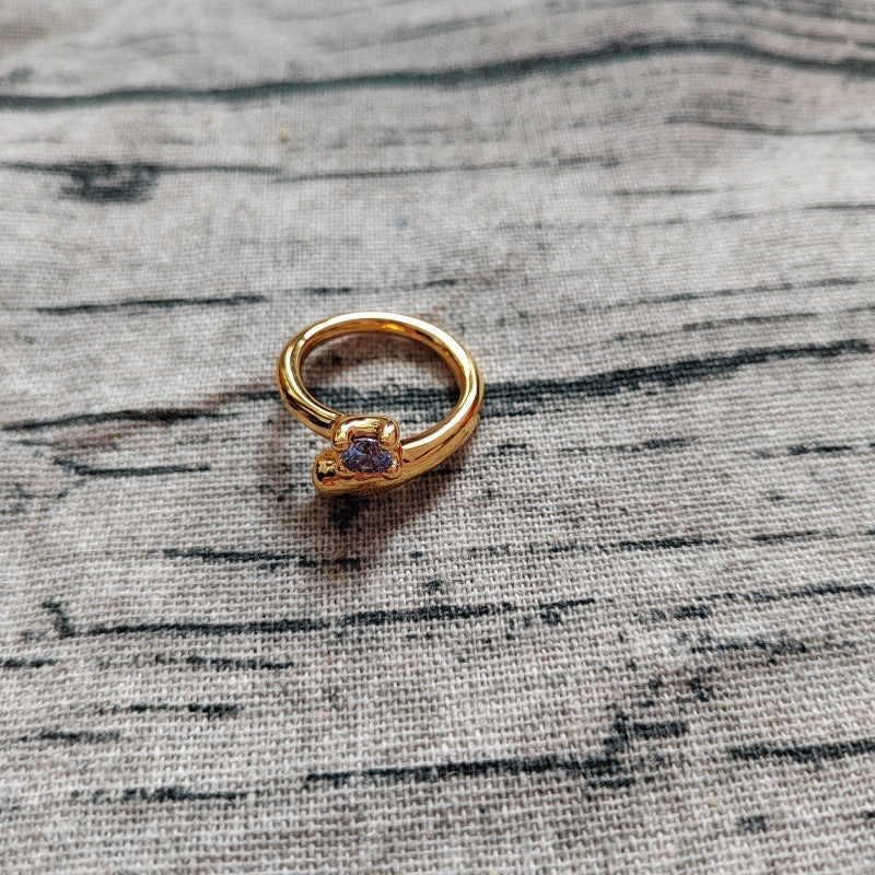 A gold ring with a small purple gemstone, resting on a textured gray linen fabric background.