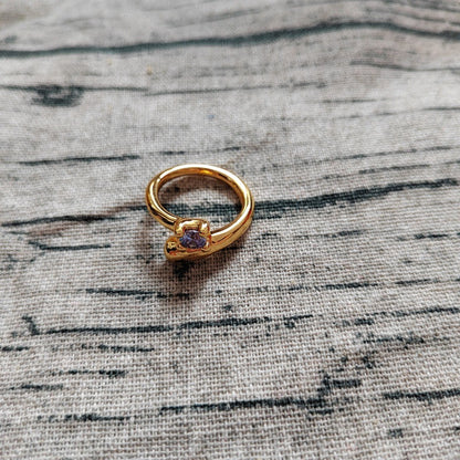 A gold ring with a small purple gemstone, resting on a textured gray linen fabric background.