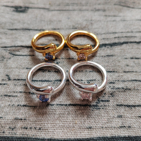 Four small rings arranged in a 2x2 grid on a textured gray fabric background. The top pair has polished gold-toned metal with a small blue gemstone, and the bottom pair has polished silver-toned metal with a small pink gemstone.