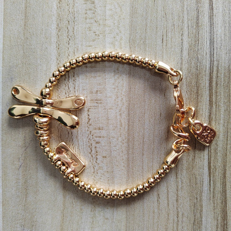 A gold-toned beaded bracelet featuring a dragonfly charm, a small rectangular charm, and a heart-shaped tag, displayed on a light wood background.