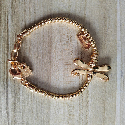 A gold-tone beaded chain bracelet with a bow charm and a small heart-shaped tag, displayed on a light wood surface.