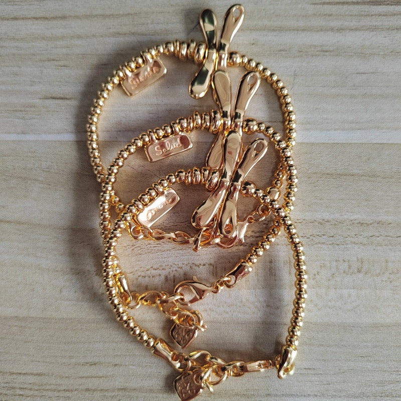Three gold-toned beaded chain bracelets with butterfly charms and heart pendants, arranged on a light wood surface.