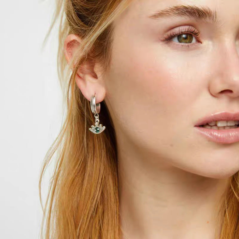 A close-up of a woman's ear wearing a silver huggie hoop earring with a dangling evil eye charm featuring green and white gemstones.