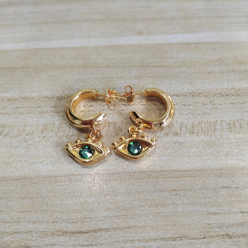 A pair of gold hoop earrings with dangling eye charms set with green faceted stones, displayed on a light wood surface.