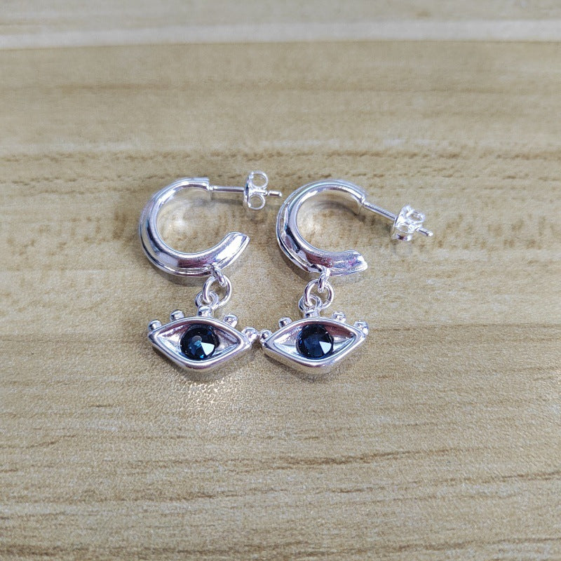 A pair of silver huggie hoop earrings with dangling eye charms featuring dark blue gemstones, displayed on a light wood grain surface.