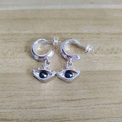 A pair of silver huggie hoop earrings with dangling eye charms featuring dark blue gemstones, displayed on a light wood grain surface.