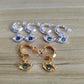 Four pairs of huggie hoop earrings with dangling eye charms are arranged on a light wood surface. Two pairs are silver-tone with dark blue-green stones, and two pairs are gold-tone with matching dark green stones.