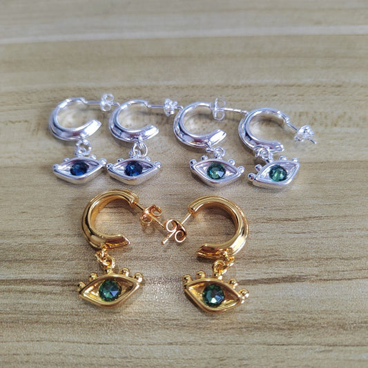 Four pairs of huggie hoop earrings with dangling eye charms are arranged on a light wood surface. Two pairs are silver-tone with dark blue-green stones, and two pairs are gold-tone with matching dark green stones.