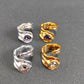 Four twisted wrap-style rings arranged on a dark gray surface, including two silver-toned rings with purple gemstones and two gold-toned rings with clear faceted gemstones.