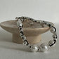 A beaded bracelet with large white pearls and polished silver-toned beads, draped over a round stone display stand against a light neutral background.
