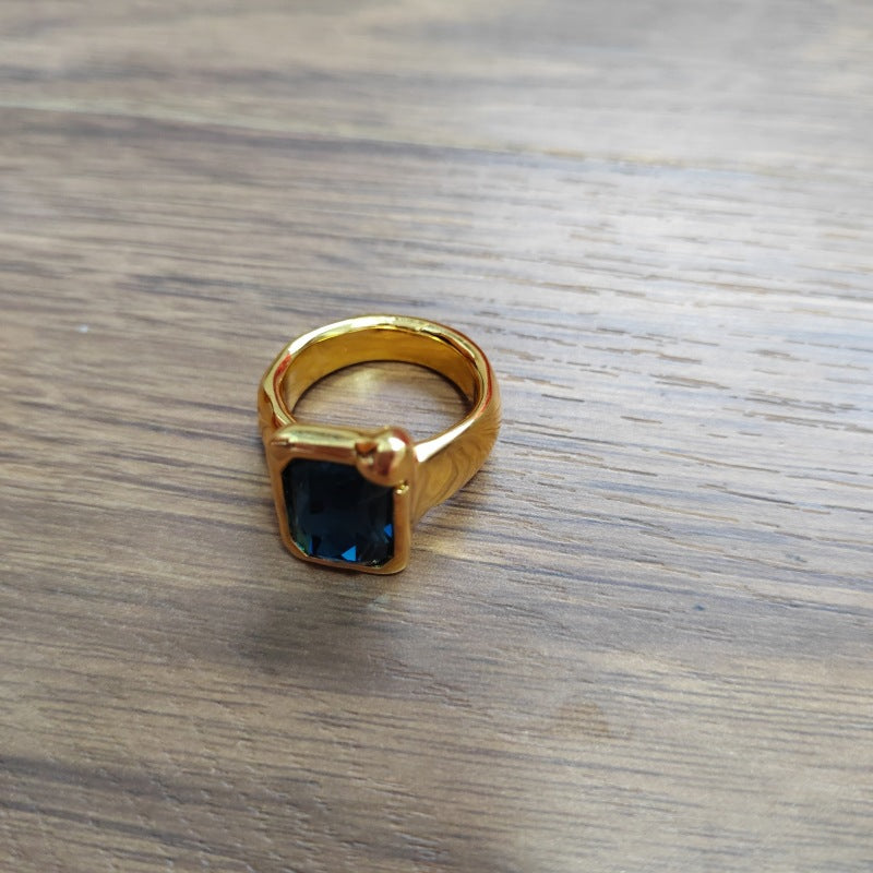 A gold signet-style ring with a square dark blue gemstone, resting on a gray wood-grain surface.