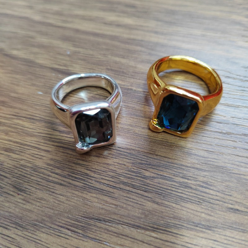 Two square-faced rings with dark blue-gray stones, one in silver-toned metal and the other in yellow gold-toned metal, displayed on a wooden surface.