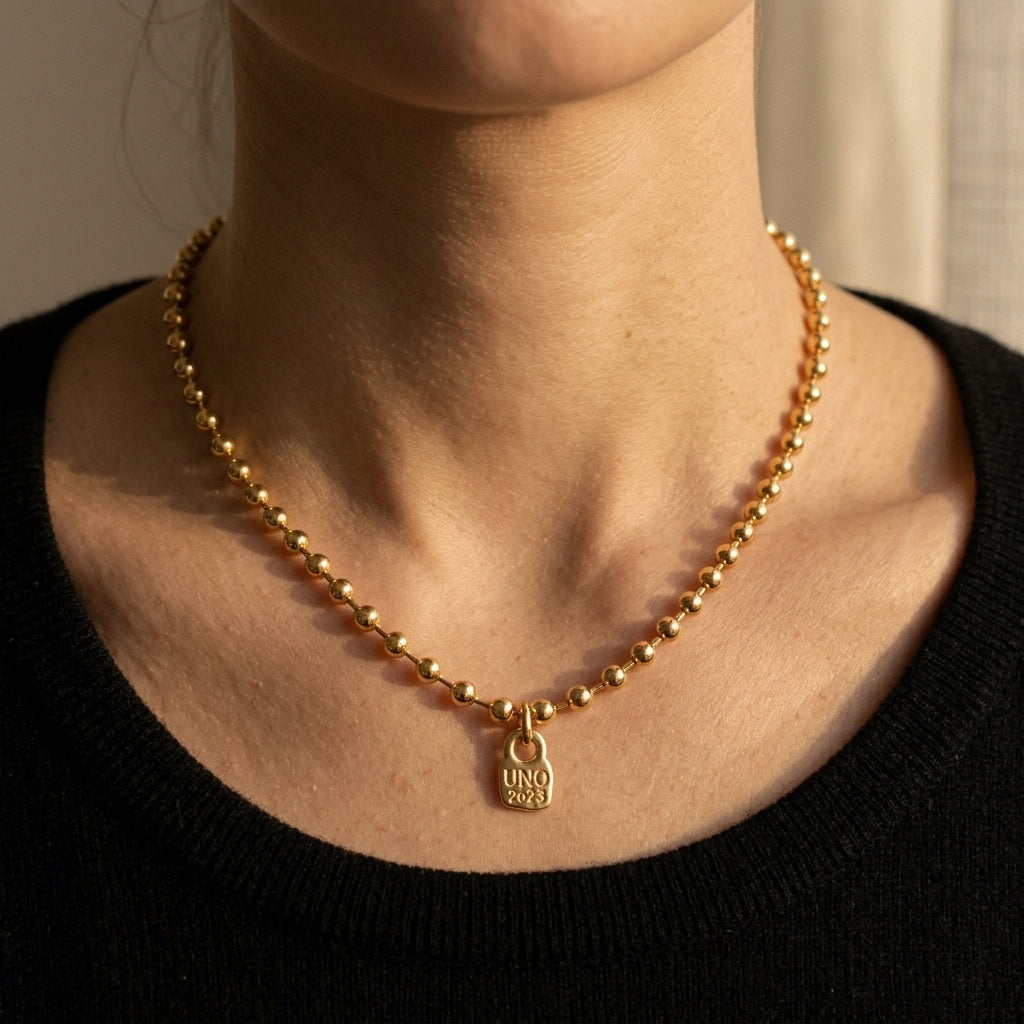 A close-up of a person's neck and upper chest wearing a delicate gold beaded necklace with a small hanging tag charm engraved with 'UNO 2025'.