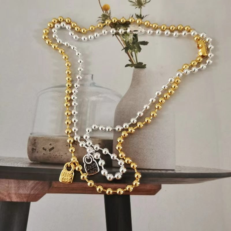 A double-strand beaded necklace with one strand of gold beads and one strand of silver beads, featuring two small hanging charms, displayed on a tabletop with a vase in the background.