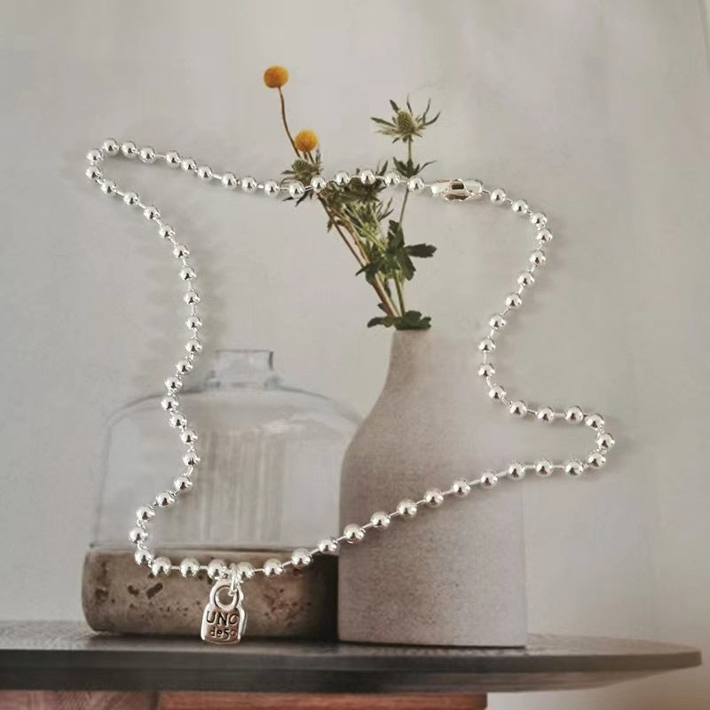 A silver ball-bead necklace with a small hanging padlock charm is draped over a tabletop setup with a clear glass bottle and a textured beige vase holding dried stems.