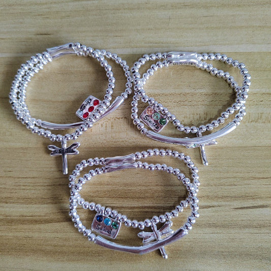 Four silver beaded bracelets with dragonfly charms and colorful gemstone accents, arranged on a light wood surface.