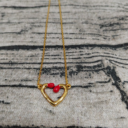 A gold necklace with an open heart pendant featuring two small red heart-shaped accents at the top, displayed on a textured gray fabric background.