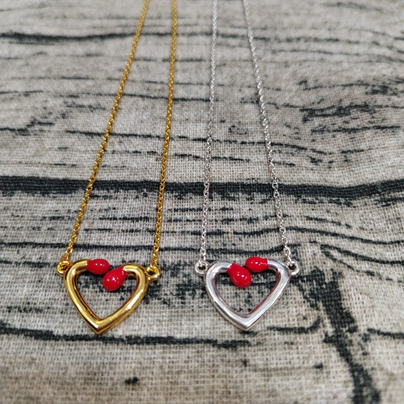 Two heart-shaped open pendant necklaces on chains, one gold-toned and one silver-toned, each with three small red heart-shaped beads at the top, displayed on a textured gray fabric background.