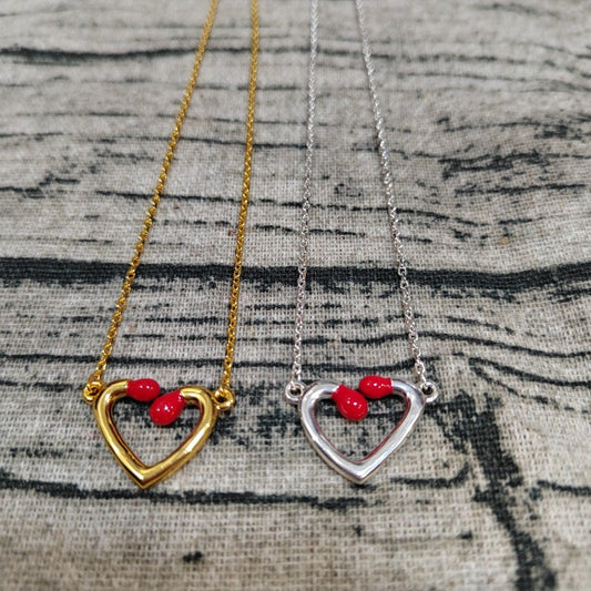Two heart-shaped open pendant necklaces on chains, one gold-toned and one silver-toned, each with three small red heart-shaped beads at the top, displayed on a textured gray fabric background.