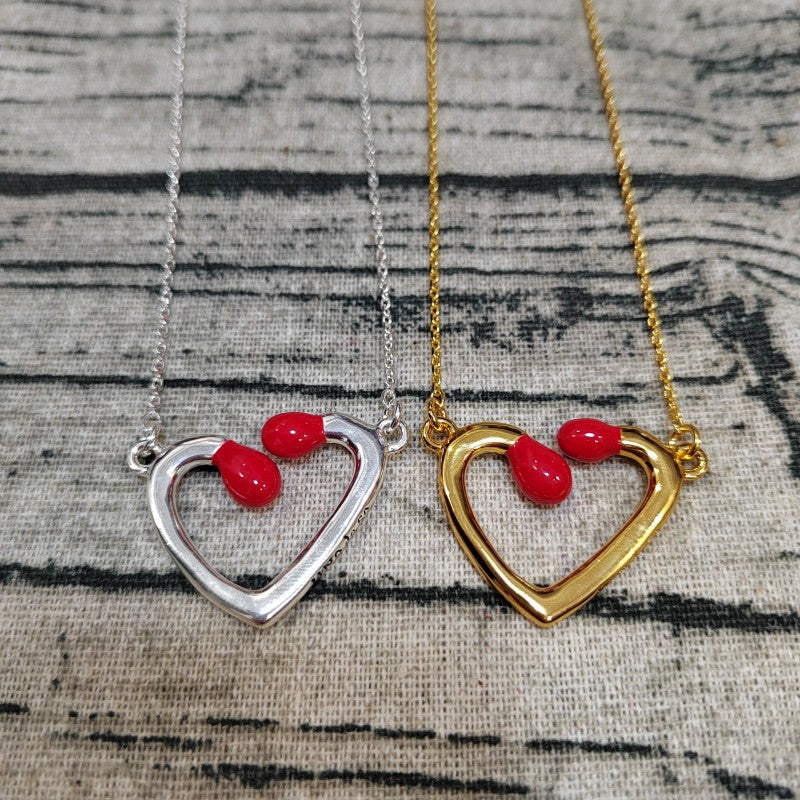 Two open heart pendant necklaces displayed side by side on a textured fabric background. One necklace has a silver-toned chain and heart, and the other has a gold-toned chain and heart, both featuring small red teardrop-shaped stones at the top of the heart.