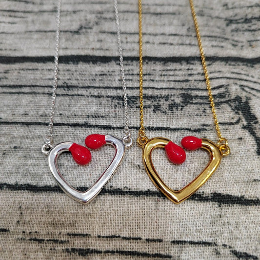 Two open heart pendant necklaces displayed side by side on a textured fabric background. One necklace has a silver-toned chain and heart, and the other has a gold-toned chain and heart, both featuring small red teardrop-shaped stones at the top of the heart.