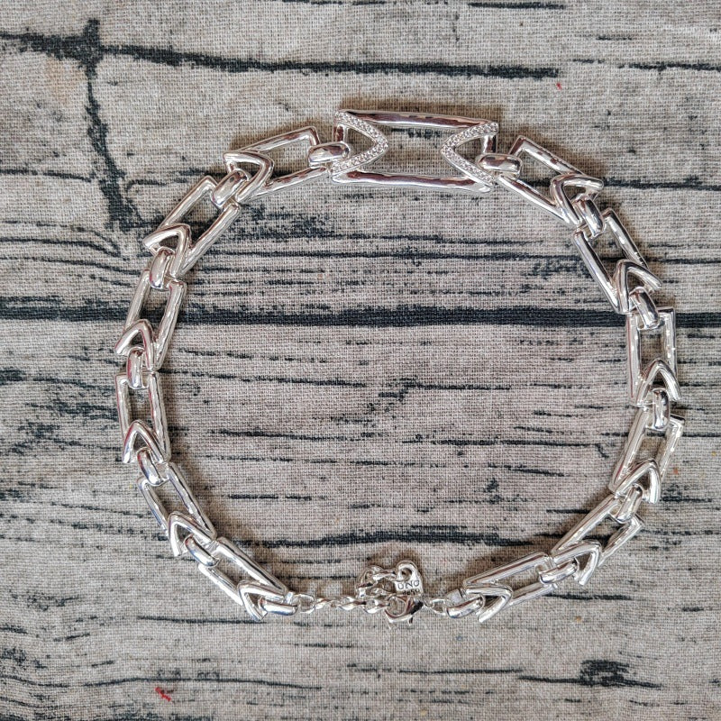 A silver-toned metal link bracelet with a geometric, interlocking chain design and a lobster clasp, displayed on a textured gray fabric background.