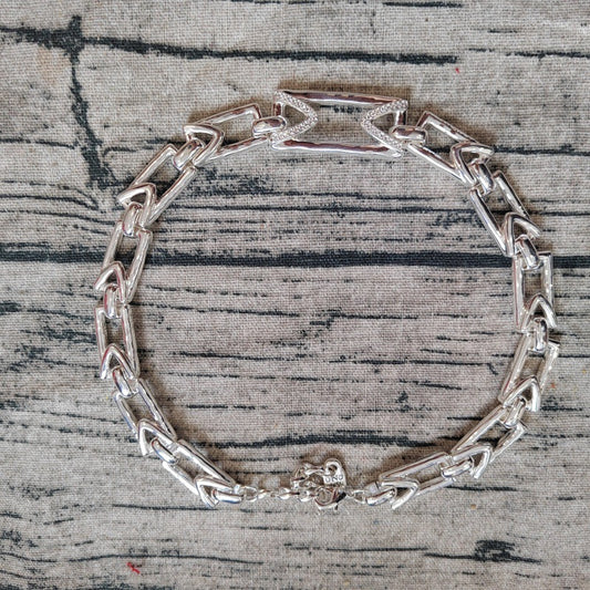 A silver-toned metal link bracelet with a geometric, interlocking chain design and a lobster clasp, displayed on a textured gray fabric background.