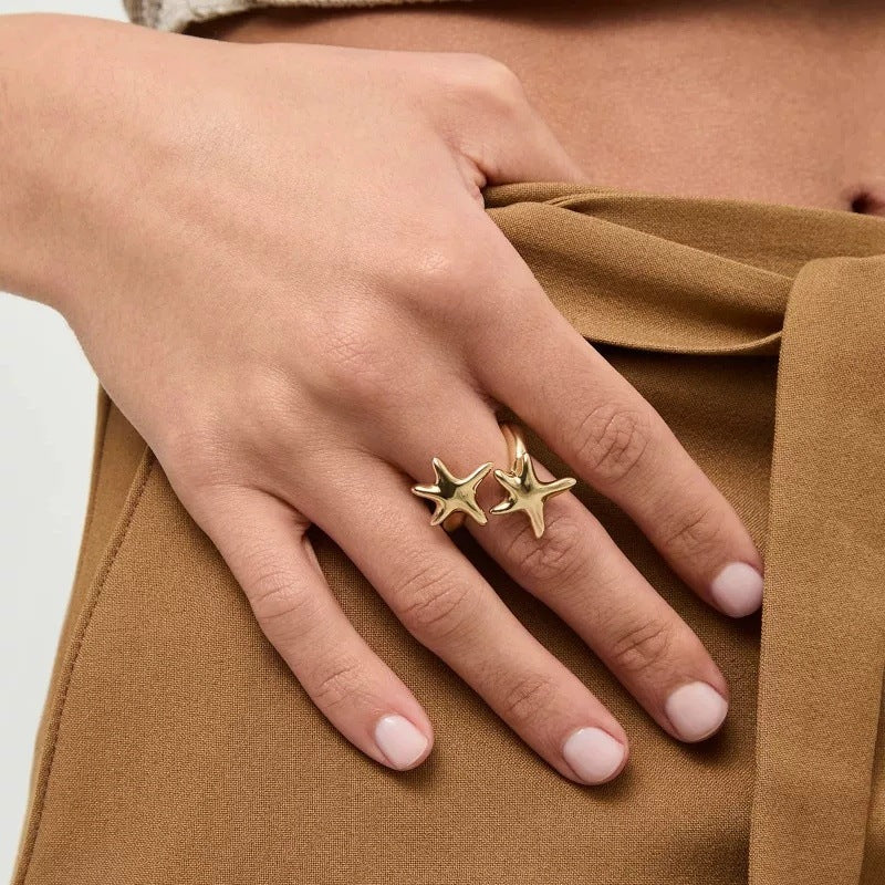A close-up of a person's hand wearing two small gold starfish rings, resting against a tan fabric surface.