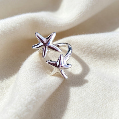A silver ring featuring two starfish motifs and a curved loop, displayed on a soft, draped cream-colored fabric.
