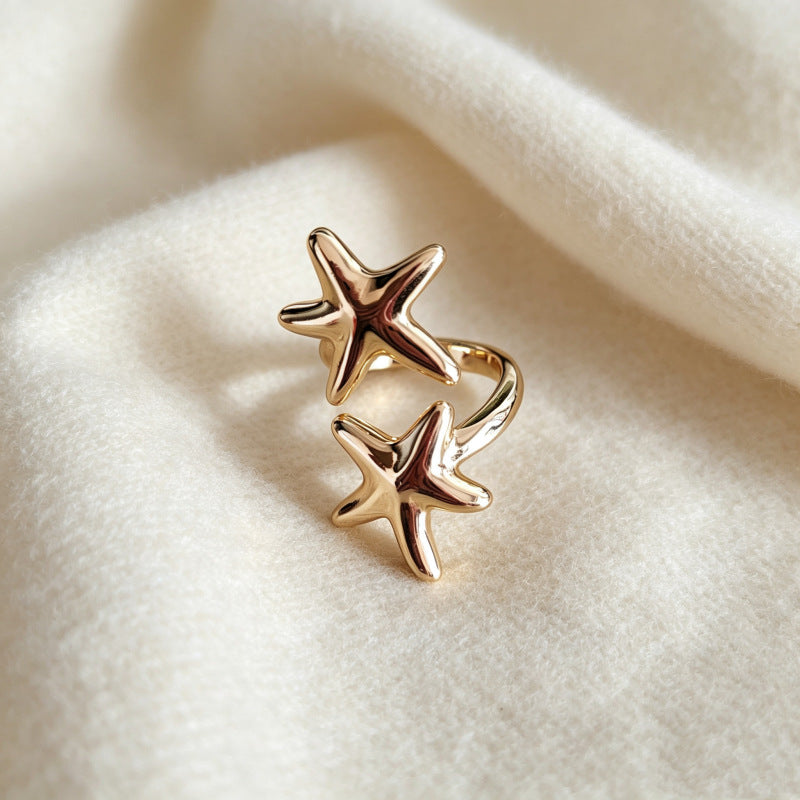 A pair of gold starfish ring charms resting on a soft, textured cream-colored fabric background.
