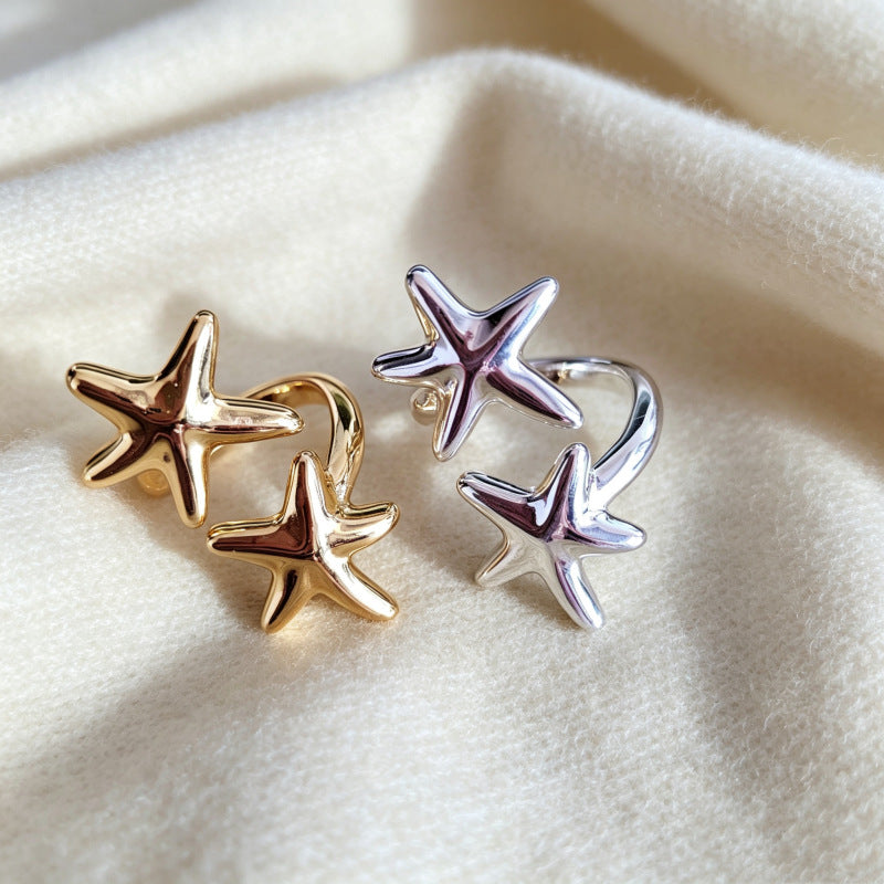 Two pairs of starfish-shaped stud earrings, one in polished gold and one in polished silver, displayed on a soft, textured cream fabric background.