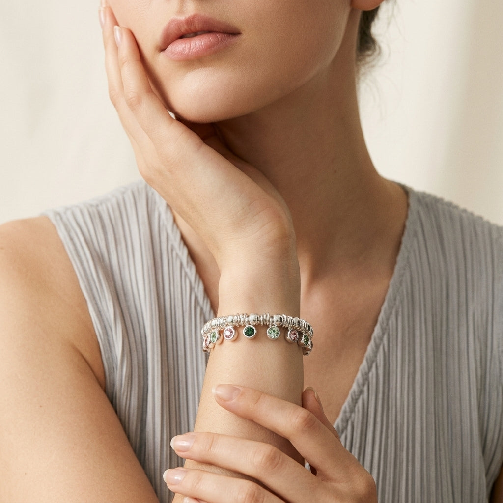 A close-up lifestyle shot of a model wearing a delicate silver-toned charm bracelet on her wrist. The bracelet features small round charms in pink, green, and clear stones.