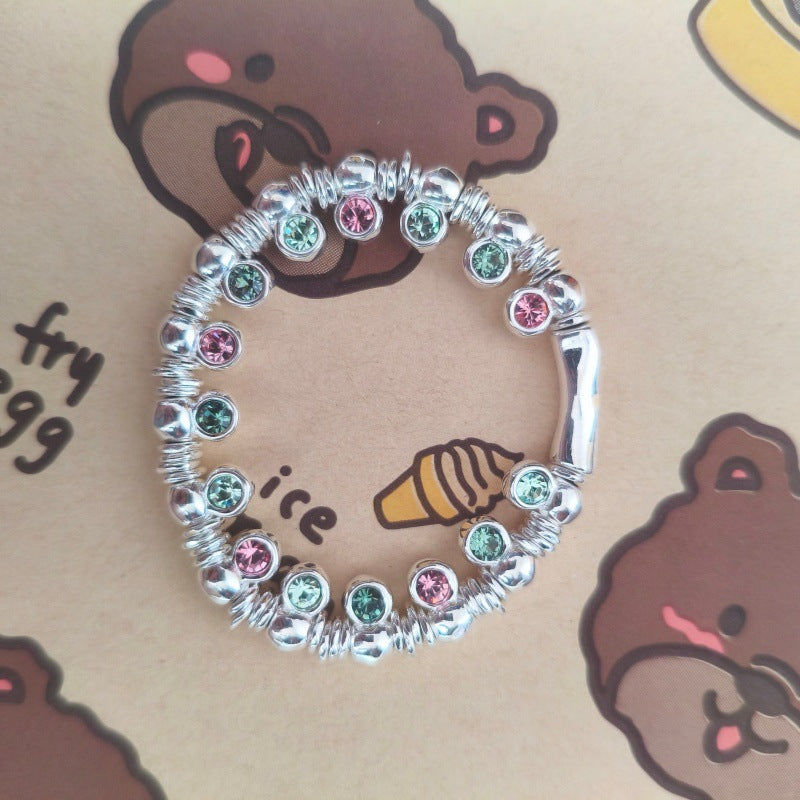 A silver-toned circular bracelet or ring embellished with small faceted beads in pink and green, photographed on a tan printed surface with cartoon bear faces and partial text.