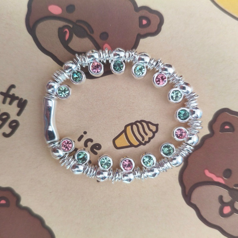 A silver-toned beaded bracelet with alternating pink and green rhinestone accents, displayed on a brown printed card featuring cartoon bear faces and an ice cream cone illustration.