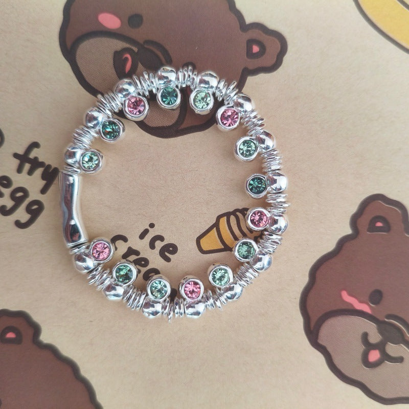 A silver-toned beaded bracelet with alternating pink and green rhinestone accents, displayed on a brown cartoon bear-patterned background.