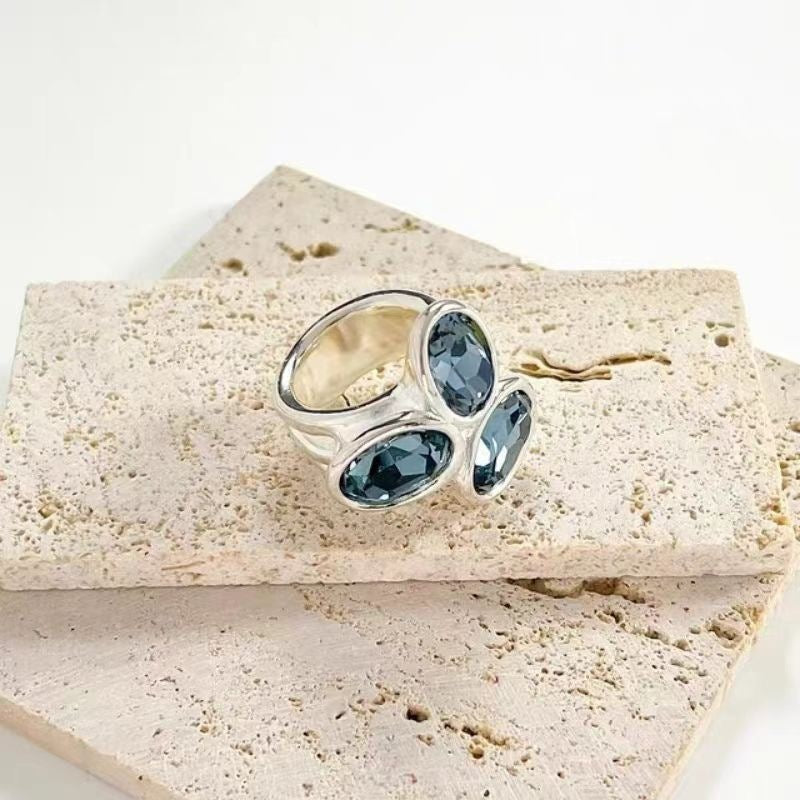 A silver ring with three marquise-cut dark blue gemstones, displayed on stacked beige travertine stone slabs against a clean white background.