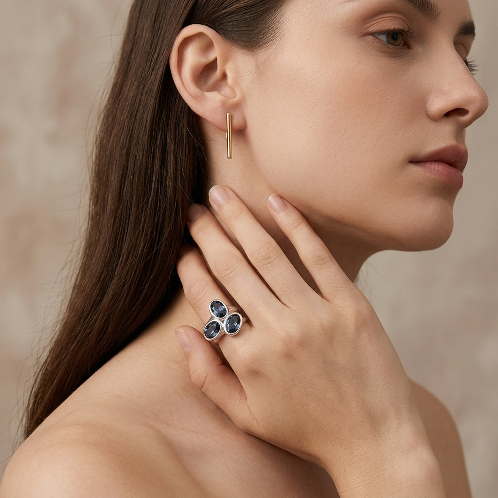 A close-up lifestyle shot of a woman wearing a silver ring with three dark blue oval gemstones and a small gold earring, set against a soft beige background.
