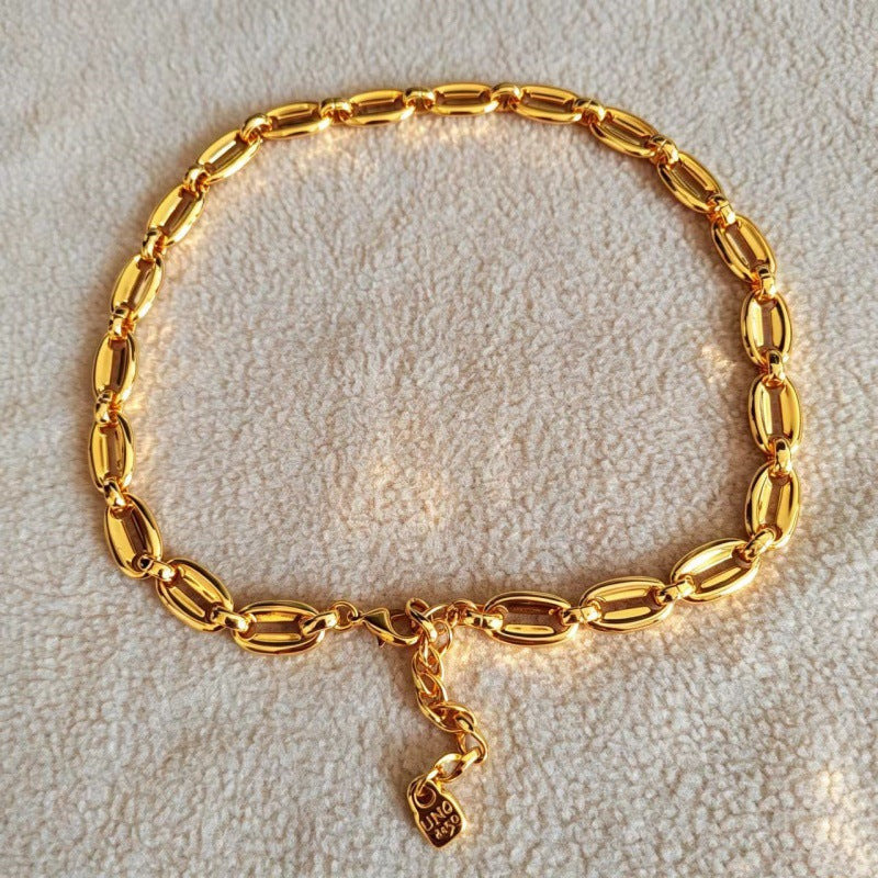 A gold chain bracelet with elongated oval links and a small hanging charm, displayed on a textured cream fabric background.