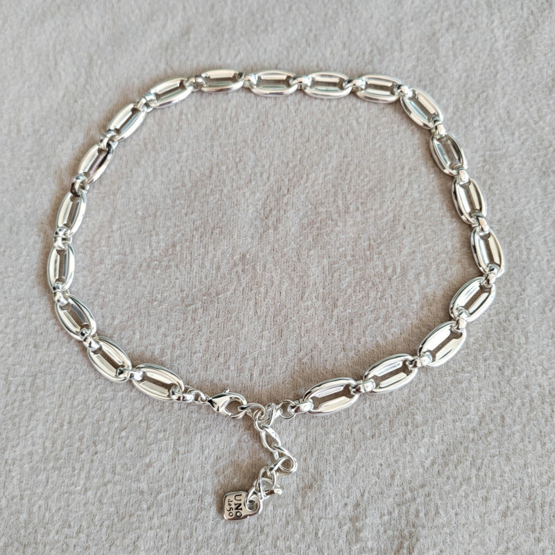 A silver link bracelet with elongated oval links and a small hanging charm, displayed on a textured light gray fabric background.