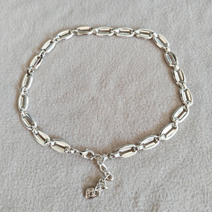 A silver link bracelet with elongated oval links and a small hanging charm, displayed on a textured light gray fabric background.