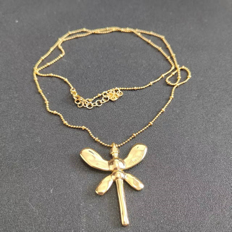 A gold dragonfly pendant necklace on a matching gold beaded chain, displayed on a dark gray textured background.