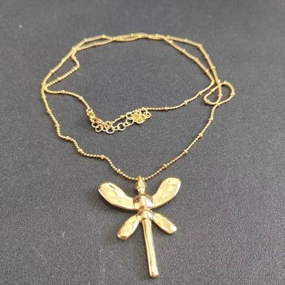 A gold dragonfly pendant necklace on a matching gold beaded chain, displayed on a dark gray textured background.