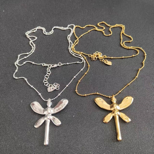 Two dragonfly necklaces displayed on a dark gray background, one in sterling silver and one in yellow gold, both with beaded chains and detailed wing pendants.