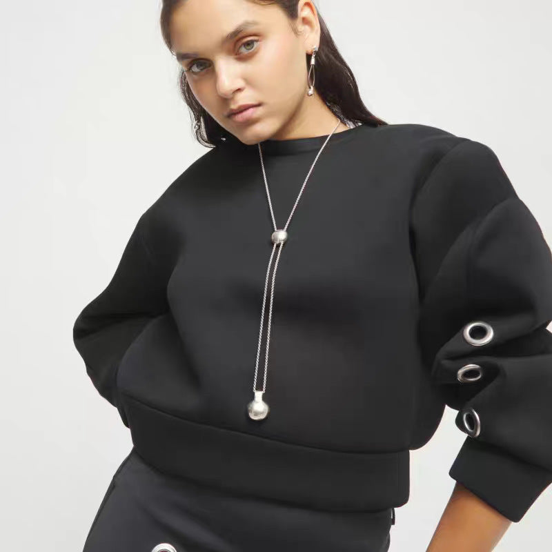 A model wearing a black crewneck sweatshirt with voluminous sleeves featuring metal grommets, paired with a long silver pendant necklace.