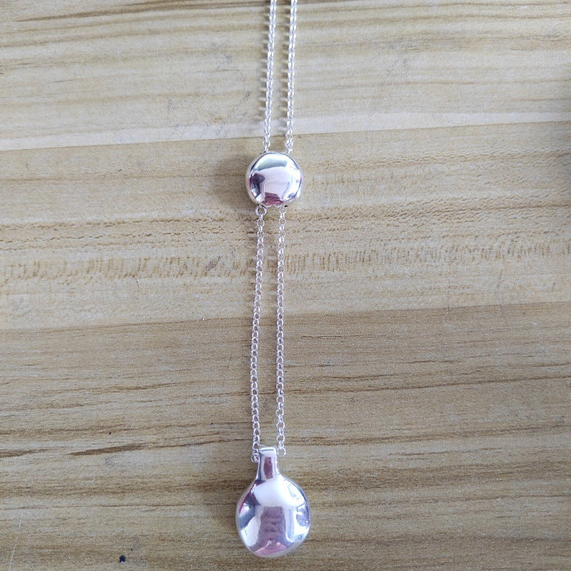 A silver-toned lariat-style necklace with a round top bead, a long chain drape, and a teardrop-shaped pendant, displayed on a light wood grain surface.