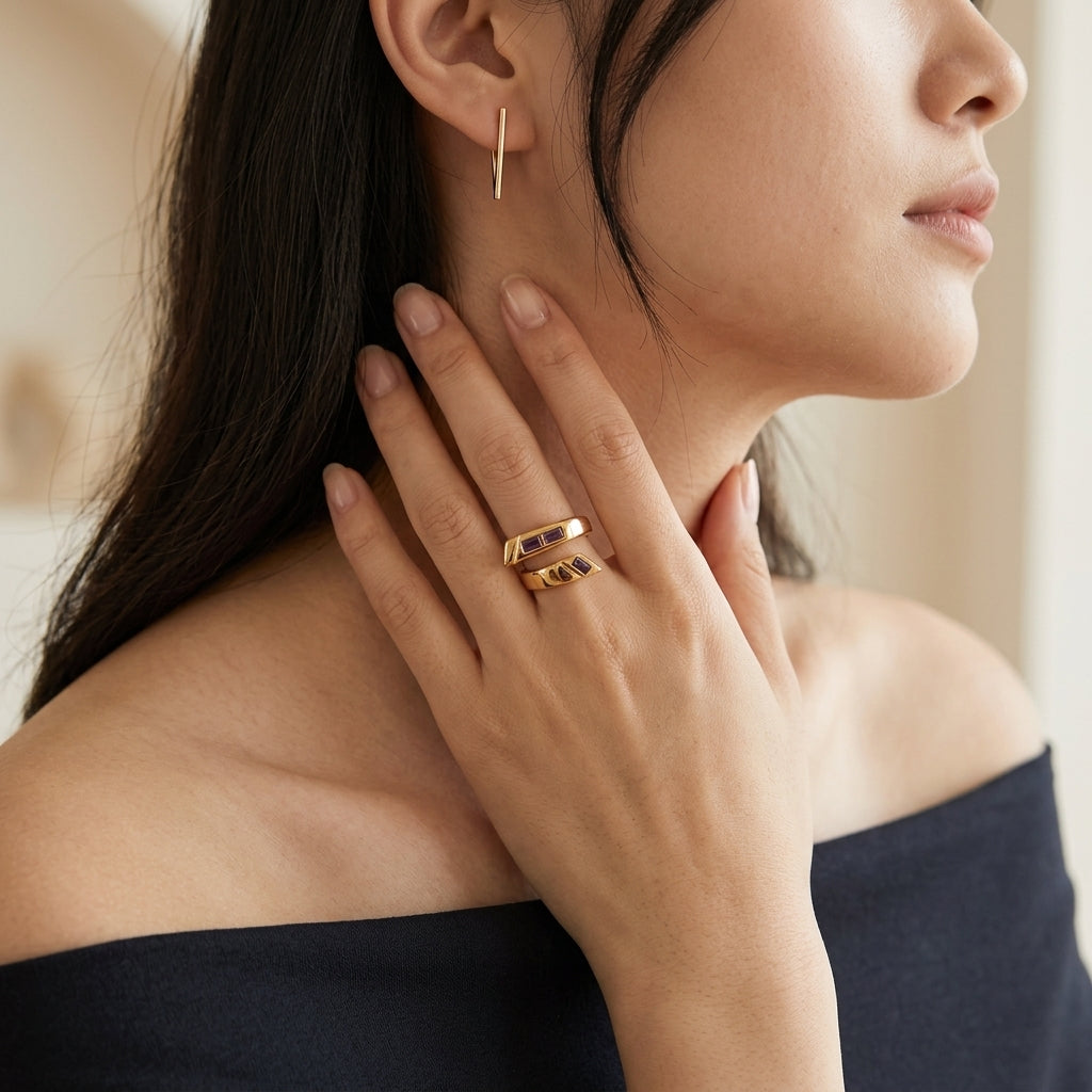 A close-up lifestyle shot of a woman wearing a double-band gold ring on her finger, with a matching small gold hoop earring visible on her ear. She is wearing a dark navy off-the-shoulder top against a soft, warm beige background.
